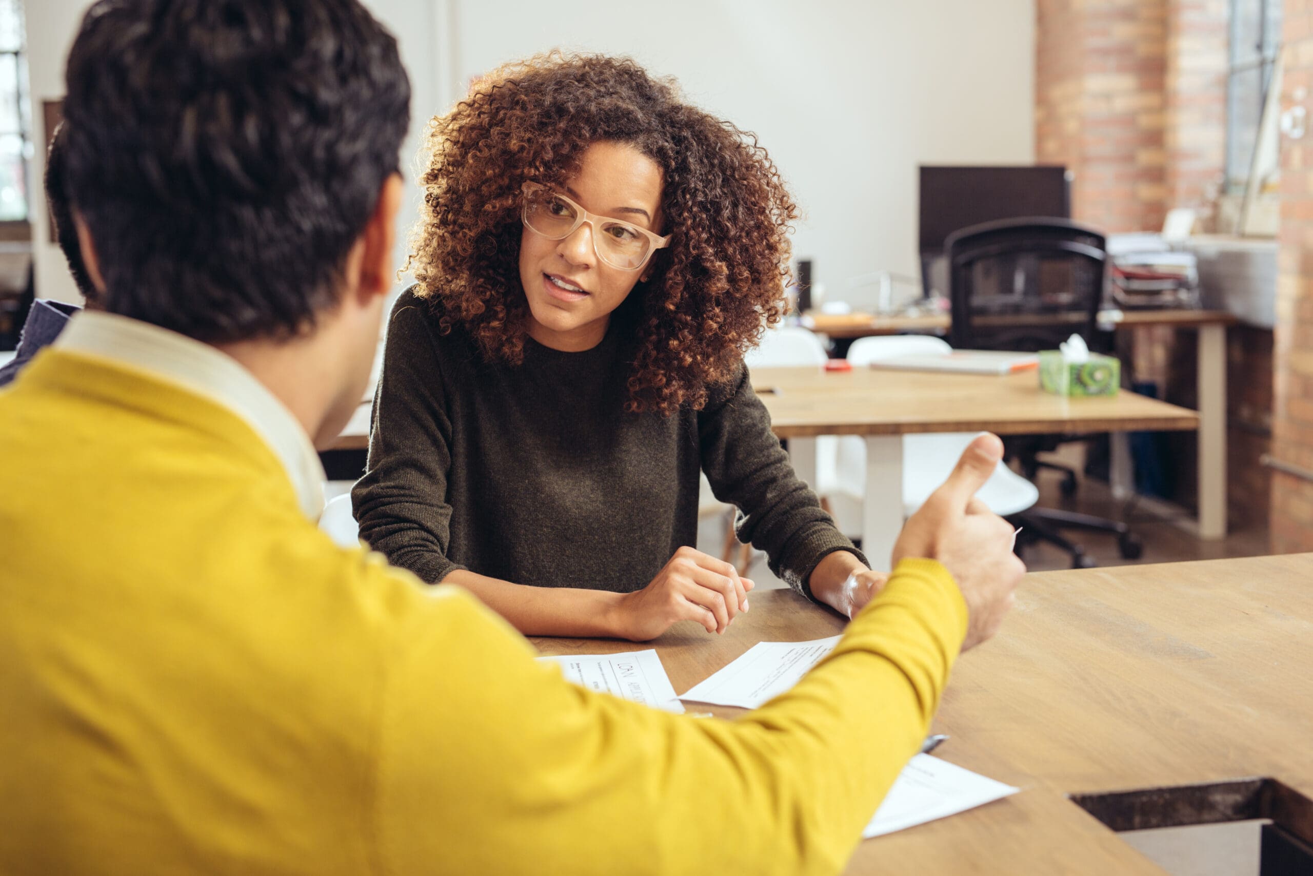 Two people sitting at a table, engaged in conversation. The person in focus has curly hair and glasses, looking towards the other person who is wearing a yellow top, holding papers.