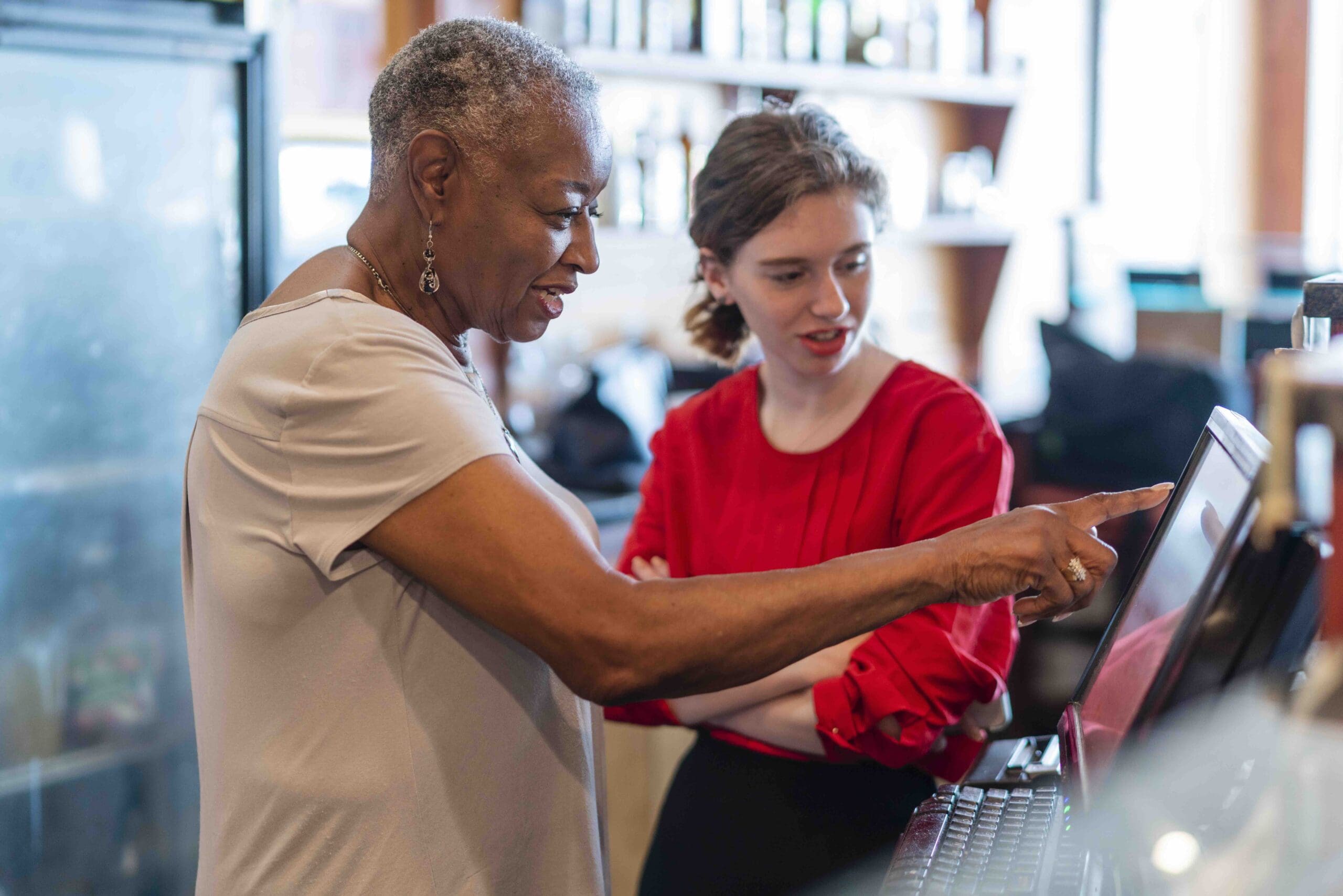 Two women stand by a computer screen. The older woman points at the screen while the younger woman watches attentively. They are in a well-lit room with shelves in the background.