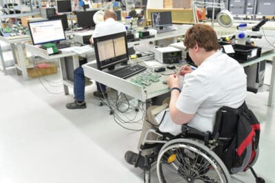 A person in a wheelchair works on electronic components at a desk in a lab, with computers and tools around. Another person is working at a nearby station.