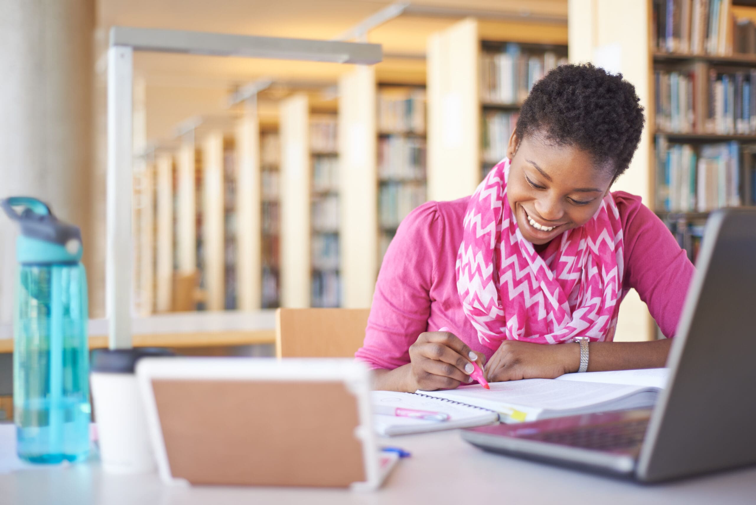 Person smiling while studying at a library table with books, a tablet, a water bottle, and a laptop.