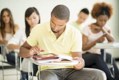 A student in a classroom focuses on a tablet and open notebook. Other students sit and concentrate in the background.
