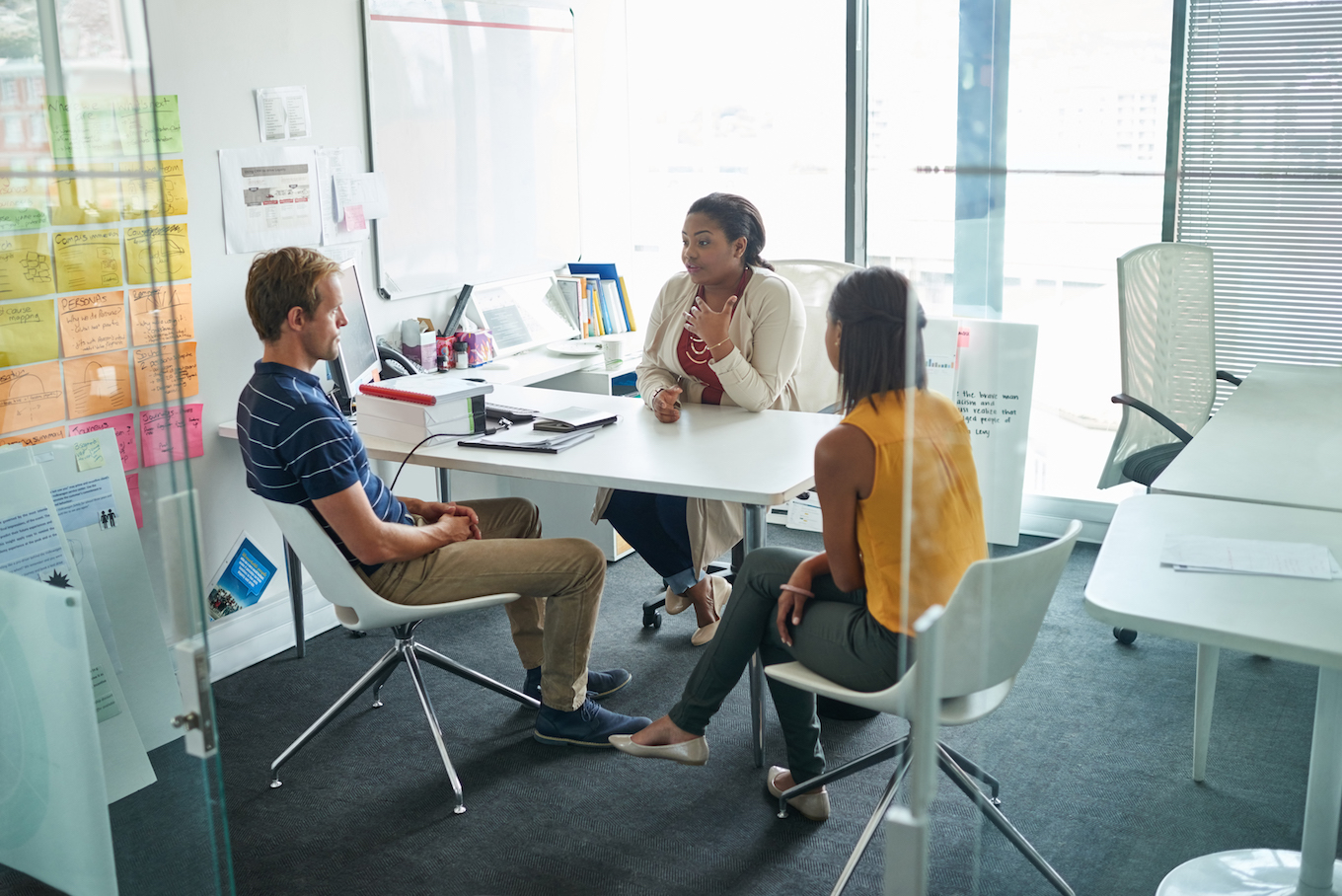 Three people having a discussion in a bright office room with post-it notes on the walls and a large window.