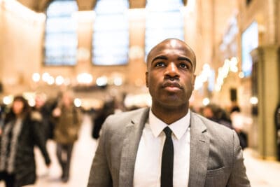 A man in a gray suit and black tie stands in a bustling, well-lit train station with blurred people in the background.