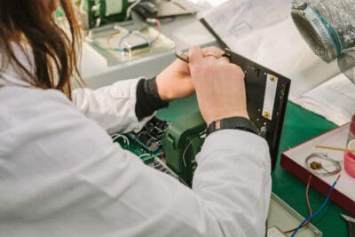 Person working on electronic components at a desk, wearing a white lab coat, using a metal tool.