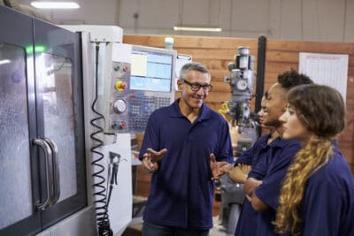 A group of people in blue shirts listen to a person explaining something near industrial machinery in a workshop.
