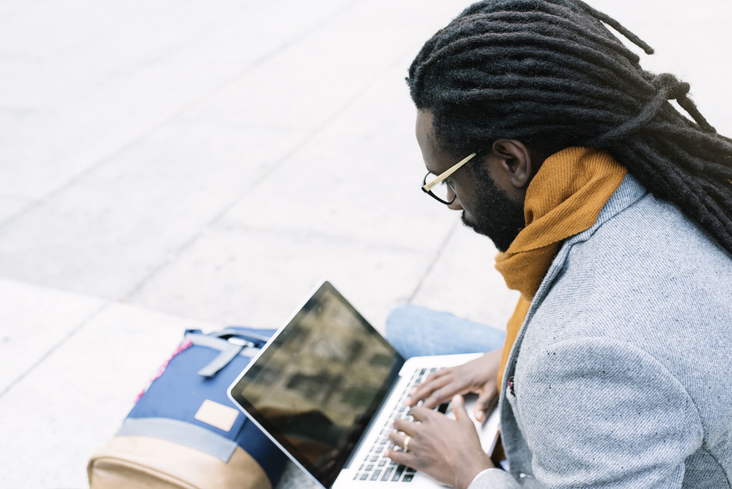 A person with long dreadlocks and glasses types on a laptop while sitting outdoors next to a backpack.