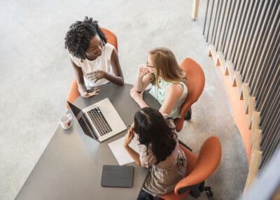 Three women are seated around a table with a laptop, papers, and a tablet. They appear to be engaged in a discussion. Orange chairs and a stripped back wall are visible.