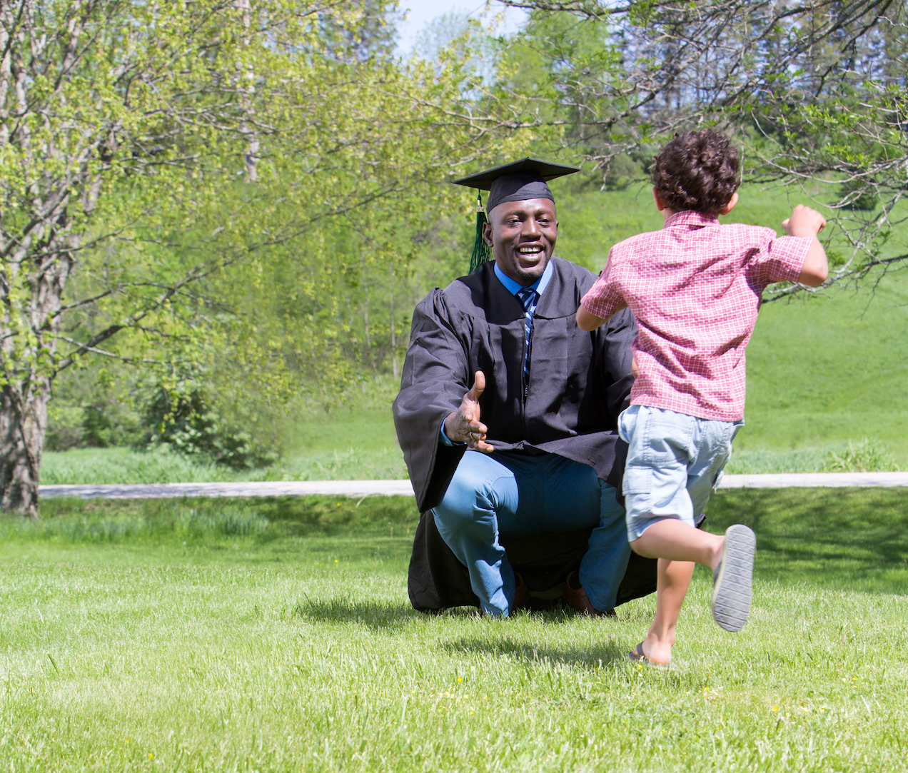 A person in graduation attire kneels on grass, arms open, as a child runs towards them in a park setting.