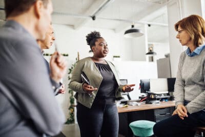 A woman is speaking to three people in an office. She is holding a tablet, while others listen attentively.