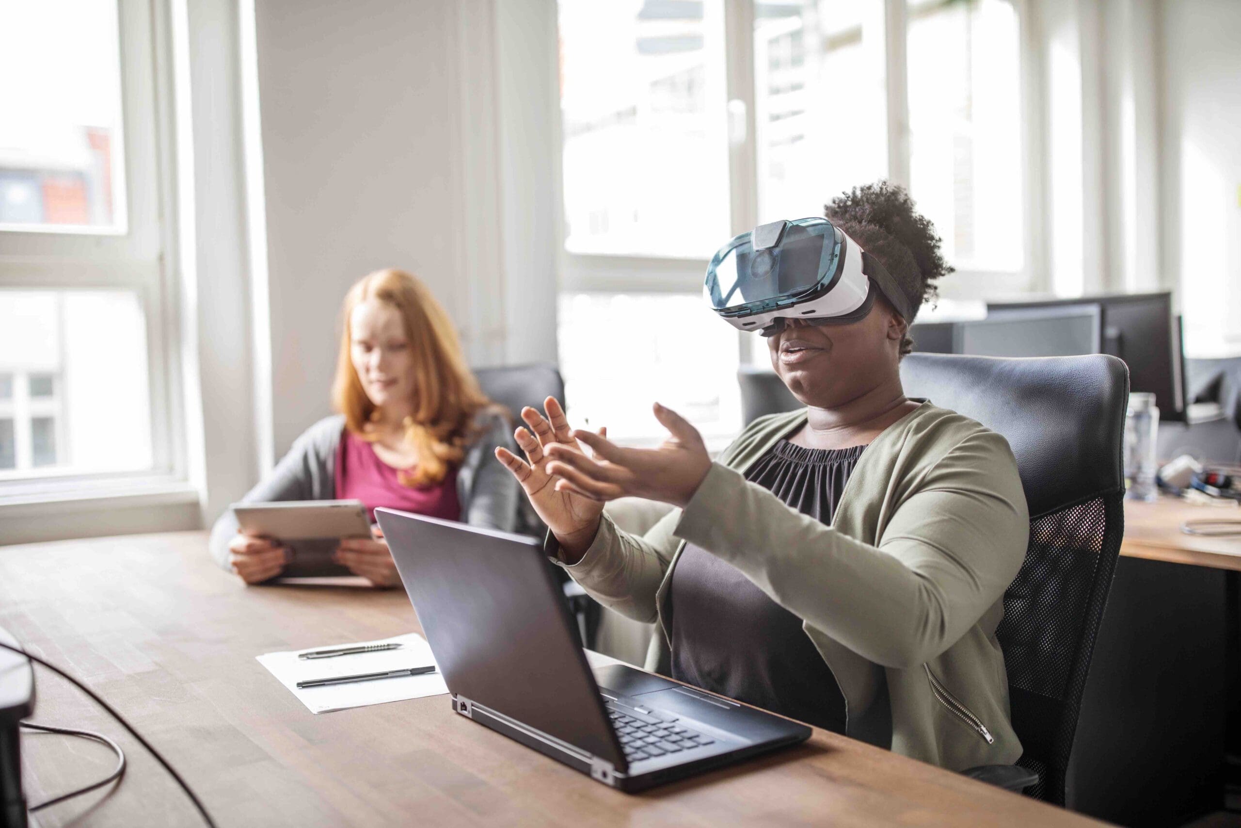 Person using a VR headset at a desk with a laptop, while another person sits nearby using a tablet.