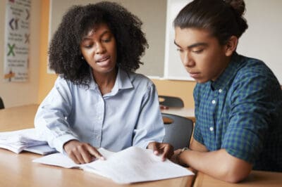 A woman and a young man are sitting at a table, looking at paperwork. The woman is pointing at the documents, engaging in discussion. Both appear focused.