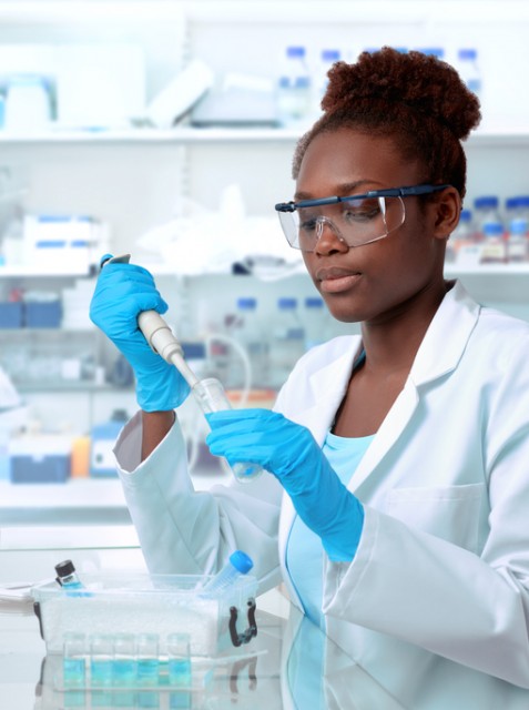 A scientist wearing goggles and gloves uses a pipette in a laboratory setting.