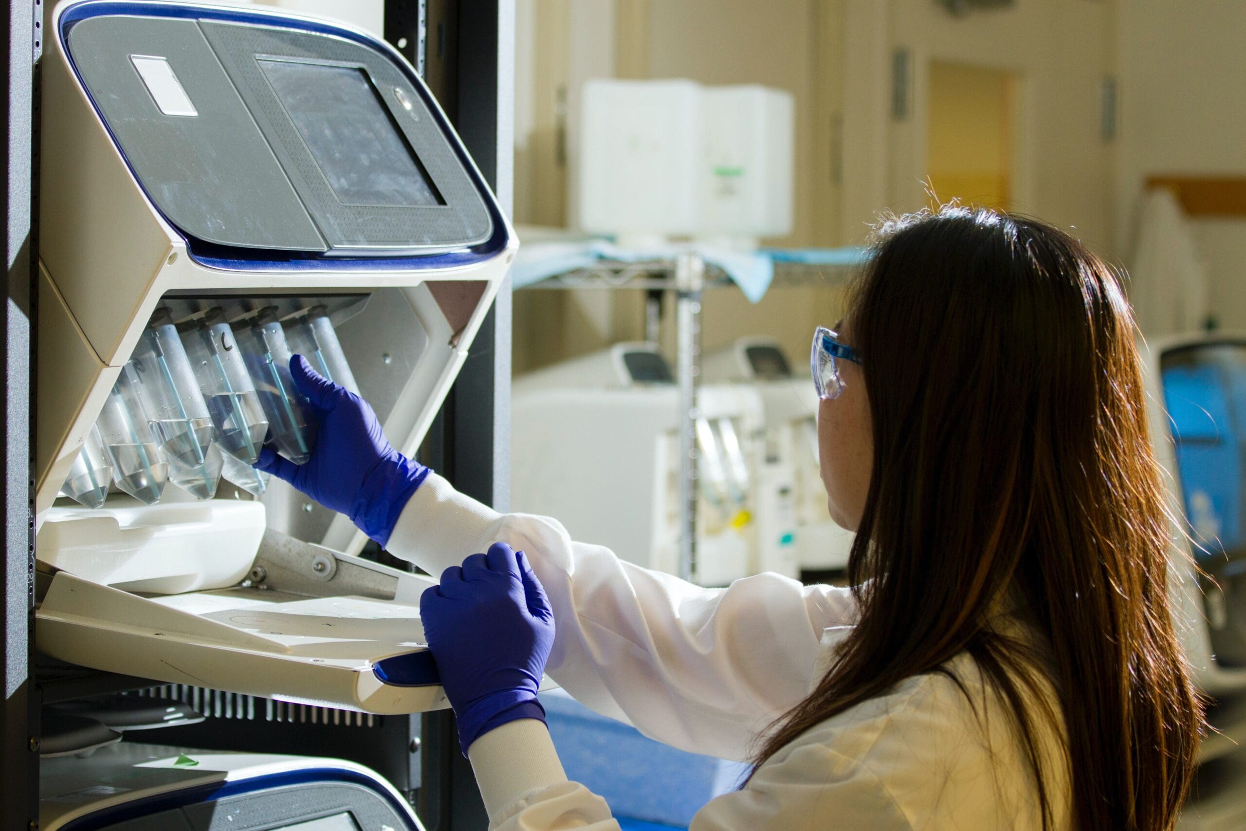 A lab technician in a white coat and blue gloves operates equipment, placing samples into a machine in a laboratory setting.