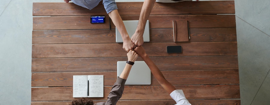 Four people fist-bumping over a wooden table with notebooks, a smartphone, and a laptop visible.