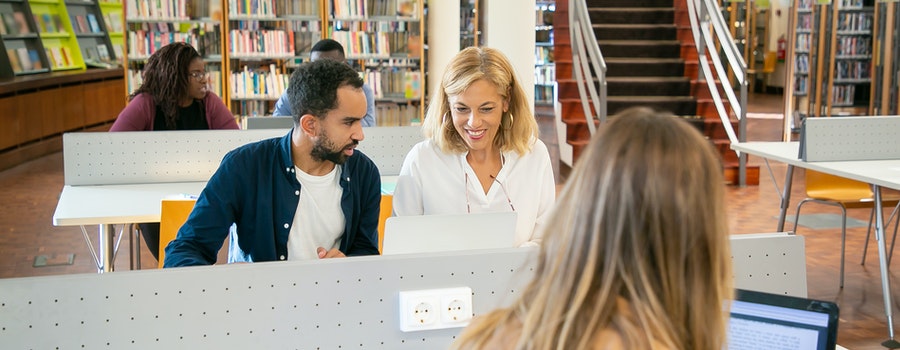 Three people working at computers in a library, engaged in conversation. Shelves of books and a staircase are visible in the background.