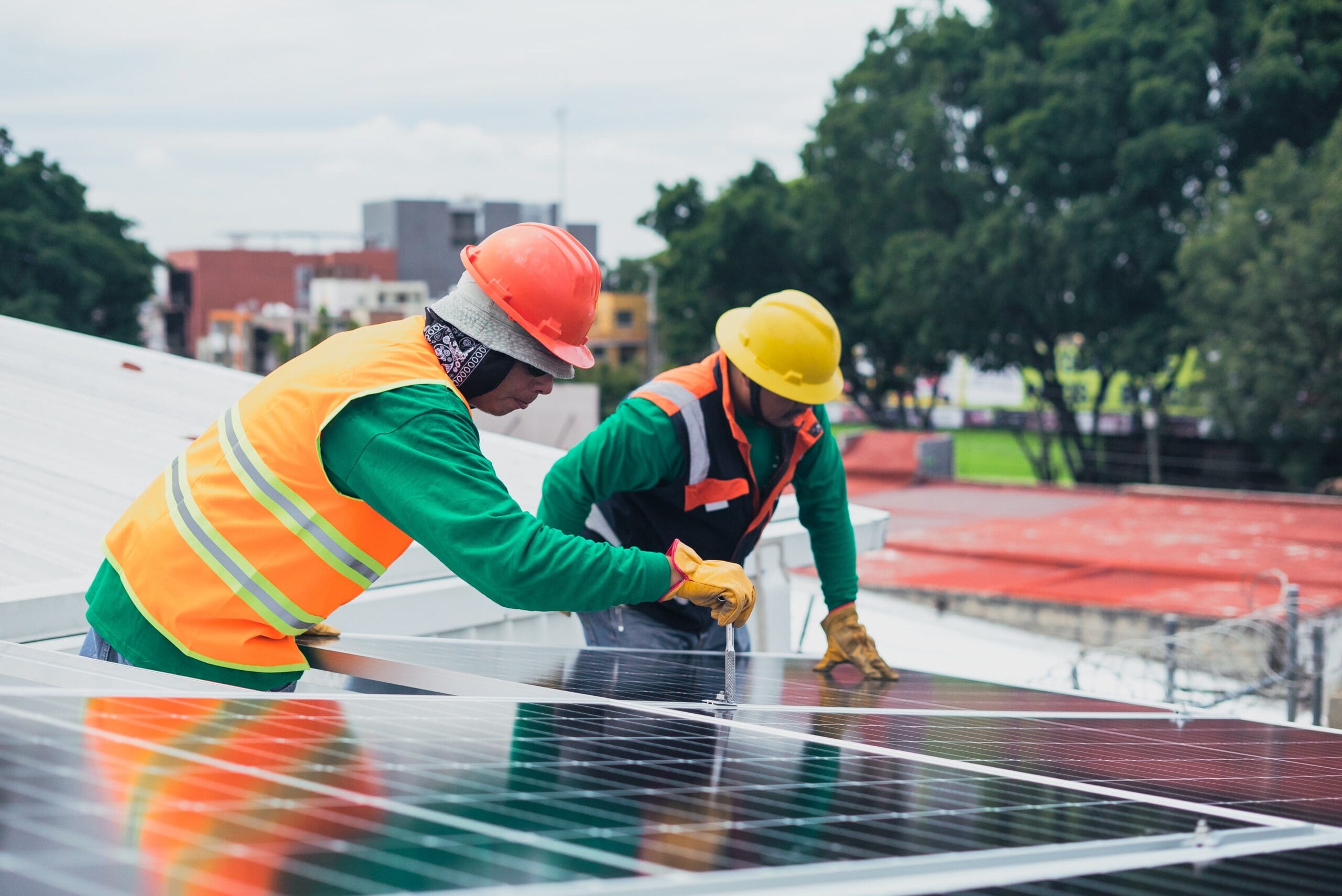 Two workers in safety gear are installing solar panels on a rooftop. Trees and buildings are visible in the background.