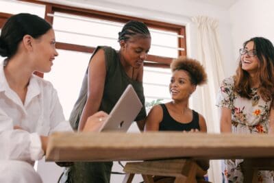 Four people are gathered around a table in discussion, with a laptop visible. One person is holding the laptop, and they appear to be engaged in a collaborative conversation.