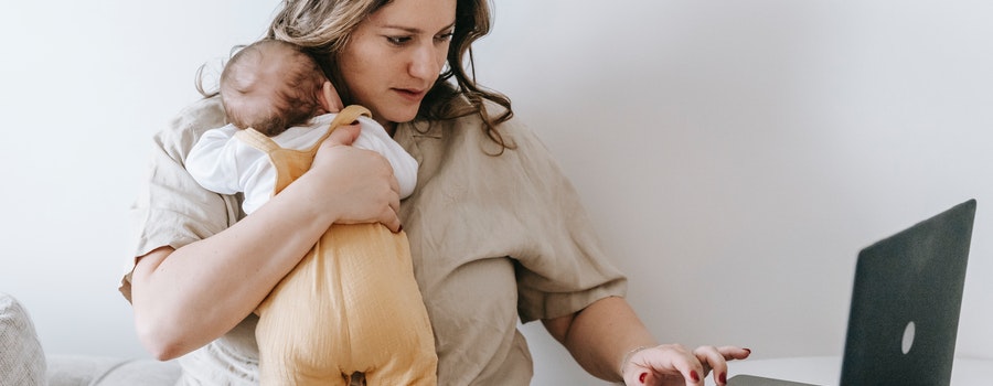A woman holds a baby while using a laptop, sitting on a couch in a white room.