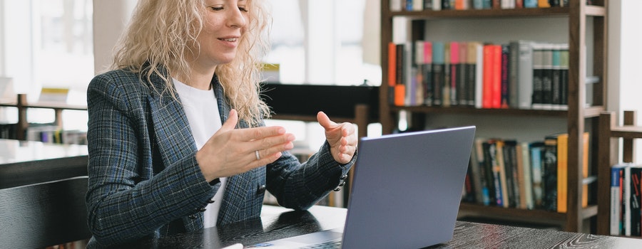 Woman sitting at a table, using a laptop, gesturing with hands, in a library setting with shelves of books in the background.