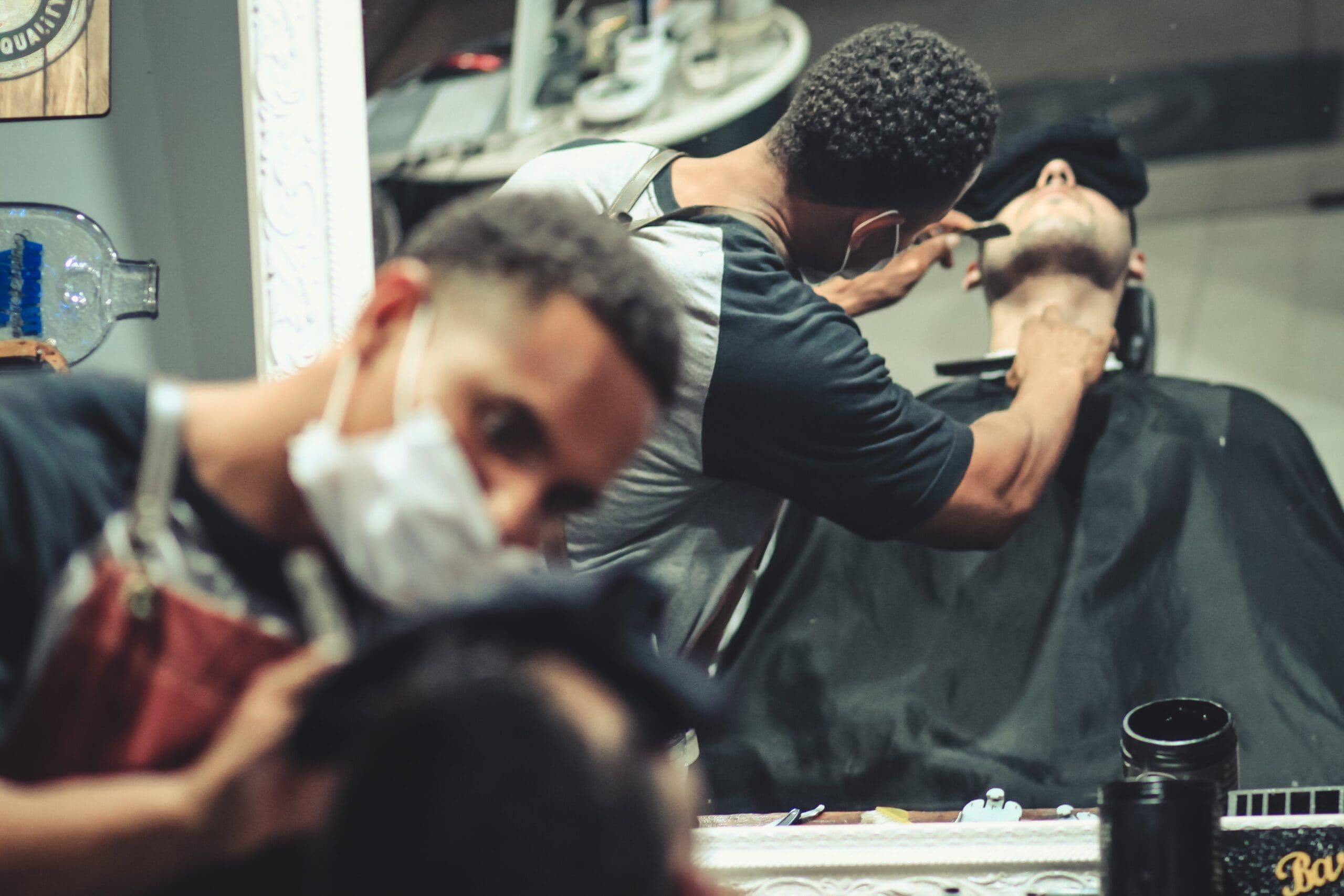 A barber trims a customer's beard while wearing a mask in front of a mirror.