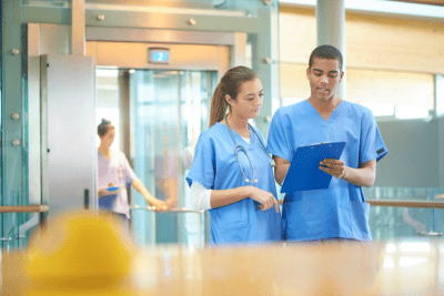 Two healthcare professionals in blue scrubs discuss notes on a clipboard in a hospital setting. A person enters through a door in the background.