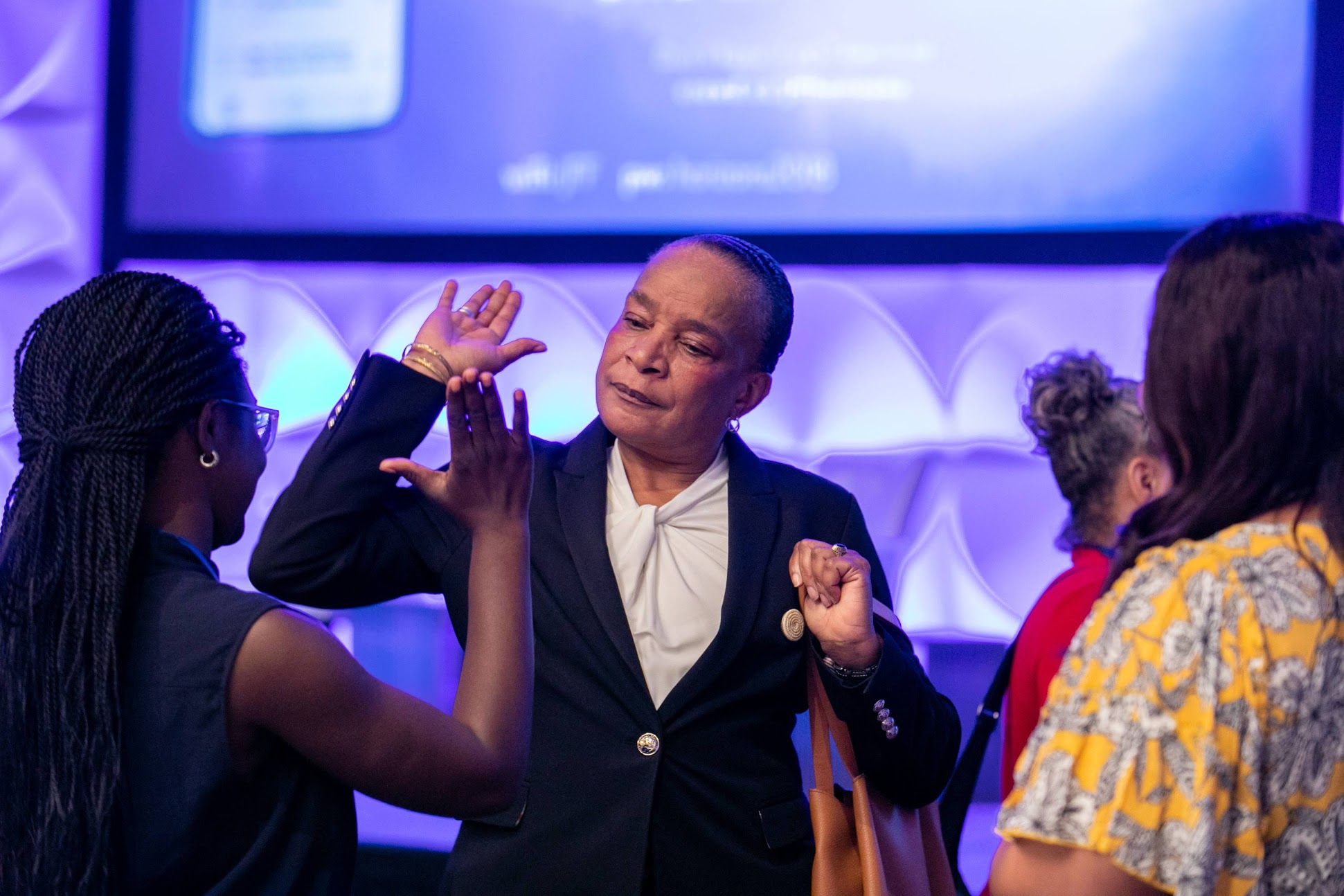 A person in a suit gestures while talking to another person in a black sleeveless top, with two more people blurred in the background under a blue-lit screen.