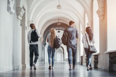 Four people walk down a corridor with arched ceilings and hanging lights, carrying backpacks.