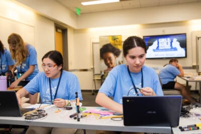Two young women work on laptops at a classroom table, surrounded by electronics and craft supplies. Others are in the background wearing similar blue shirts.
