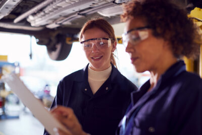 Two women in safety glasses and coveralls stand under a vehicle, reviewing a clipboard together in an automotive workshop.