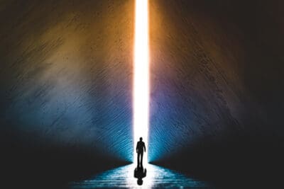 The silhouette of a man walking towards a bright light in the opened huge wall. A light in the end of a tunnel.