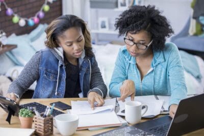 Two people work together at a table with books, papers, a laptop, and coffee cups in a cozy room.