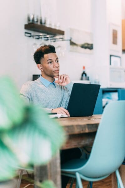 A person sits at a wooden table with a laptop, appearing thoughtful. A plant in the foreground and shelves with decorative items are visible in the background.