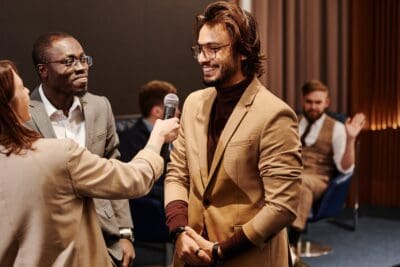 A man in a brown suit, embodying climate resilience, is holding a microphone and smiling at a woman handing it to him. Another man in a gray suit stands nearby, while in the background, a seated man is clapping.