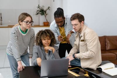Four people gather around a laptop, discussing something on the screen, in a modern office with a sofa and plants in the background.