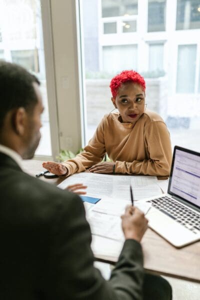 Two people sit at a table with documents and a laptop, engaged in conversation. The person facing away wears a suit, while the other person with short red hair listens attentively.