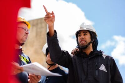 A man wearing a safety helmet points upward while speaking to colleagues, who are also wearing helmets, against a bright sky backdrop.