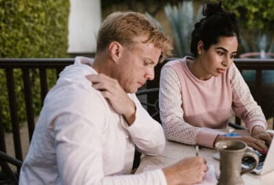 Two people write in notebook and use laptop at outdoor table