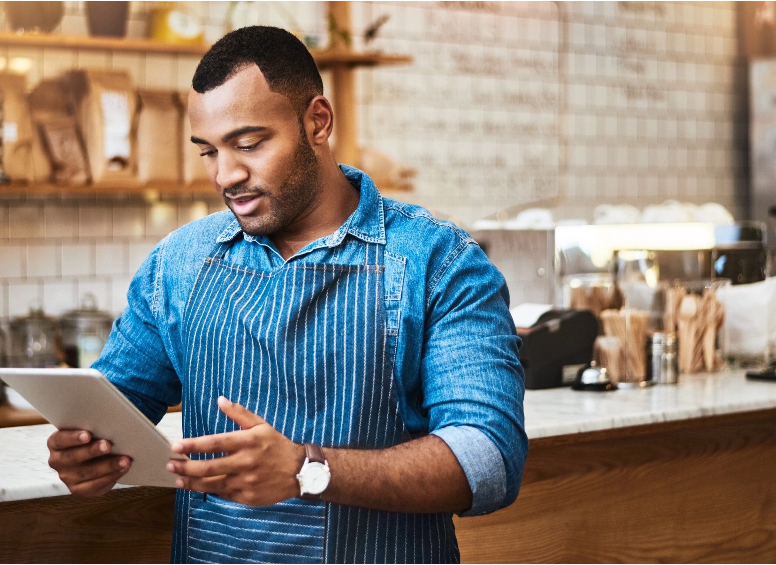 Man in a blue apron using a tablet in a cafe kitchen.