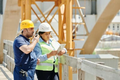Two construction workers in helmets and vests stand on a bridge, discussing the project. One reads a tablet displaying credential transparency data while the other gestures animatedly. Machinery hums in the background, underscoring their focus and expertise.
