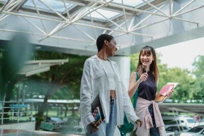 Two people walking outdoors under a glass roof, smiling and holding notebooks.