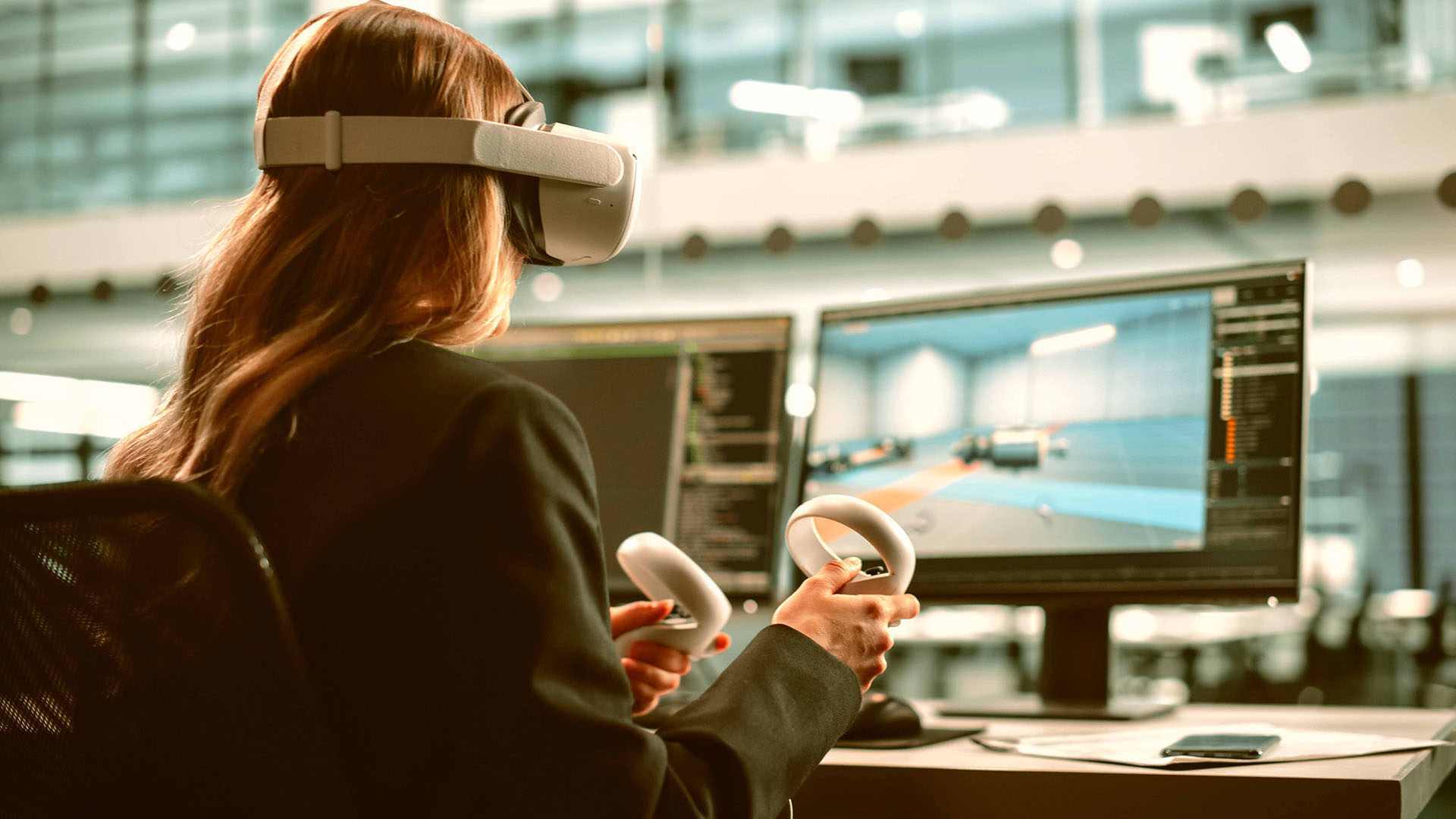 Person using VR headset and controllers, sitting at a desk with dual monitors displaying a virtual environment.