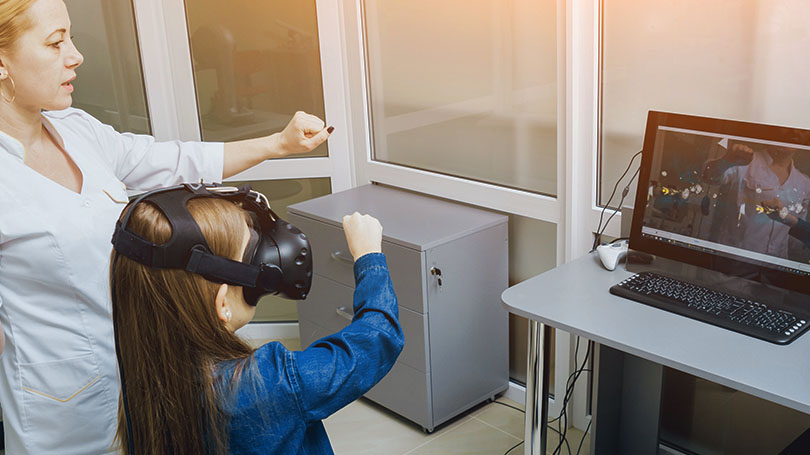 A child wearing a VR headset interacts with a virtual environment, guided by an adult in a room with a computer displaying the same scene.