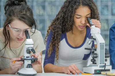 Two young women are using microscopes at a table, concentrating on their work.