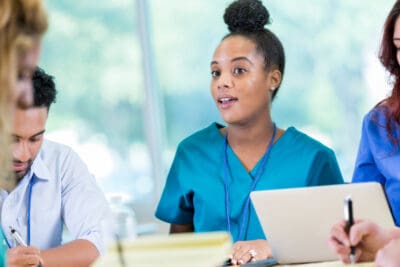 A person in a blue medical uniform speaks at a meeting. Others take notes on paper and a laptop.