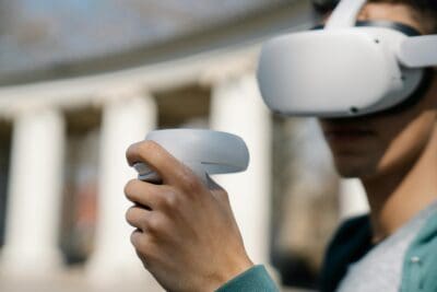 Person wearing a virtual reality headset and holding a VR controller, standing outdoors near white columns.