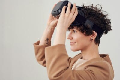 A person with short curly hair is smiling while adjusting a virtual reality headset on their head. They are wearing a beige jacket and have a small tattoo on their forearm.