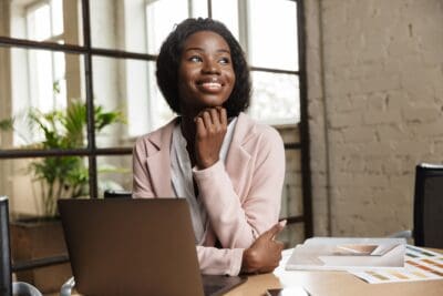 A woman in a pink blazer sits at a table with a laptop and documents, smiling and looking to the side, in a well-lit office space.