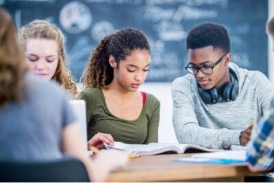 A group of students sitting at a table, focusing on textbooks in a classroom with a chalkboard in the background.
