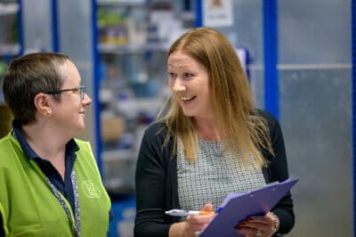 Two people engaging in a conversation inside a store. One holds a clipboard with papers. Shelves in the background contain various items.