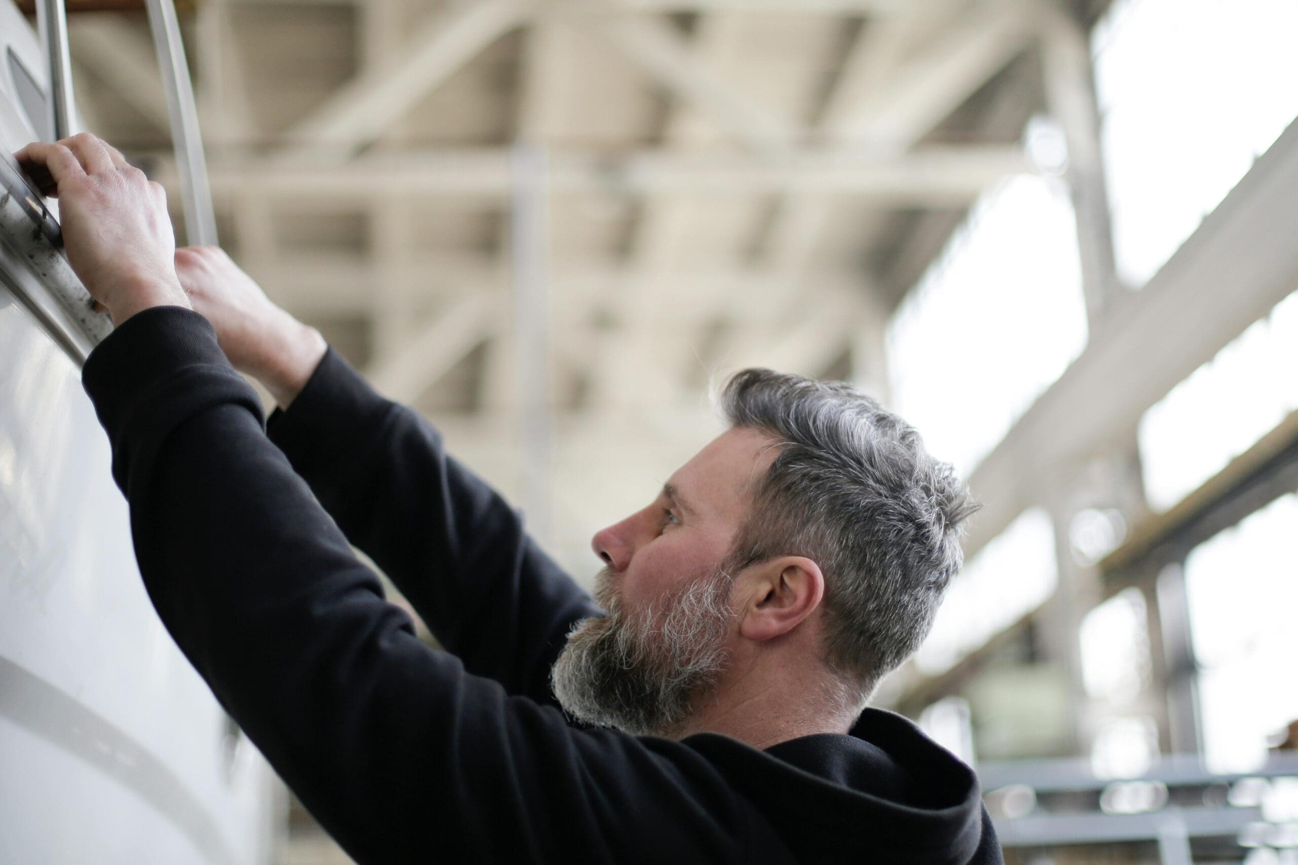 A man with a beard and gray hair works on a large white surface in a well-lit industrial setting.